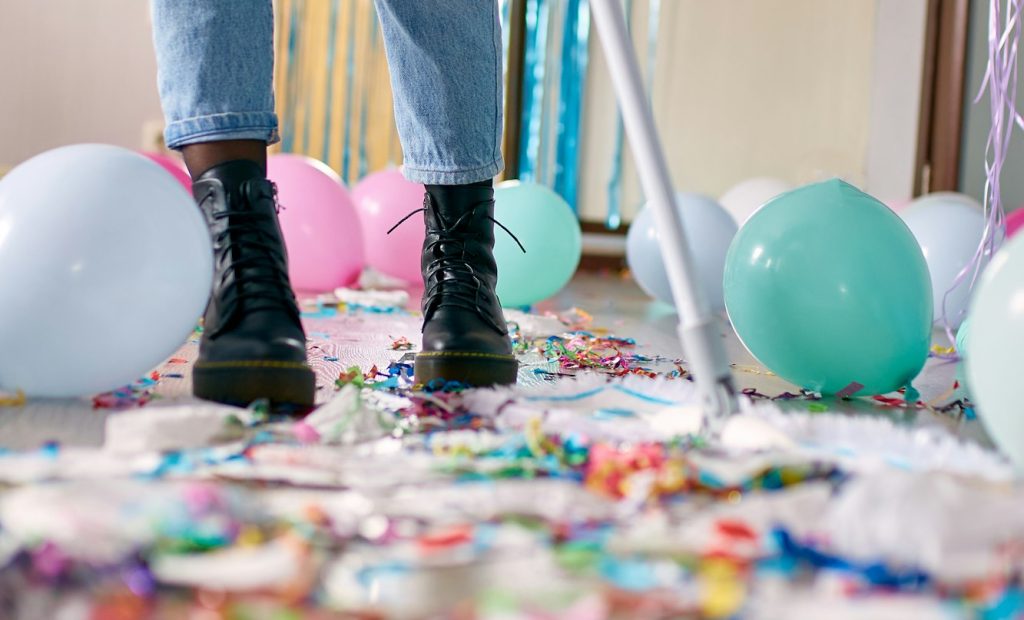 Woman with pushbroom cleaning mess of floor in room after party confetti